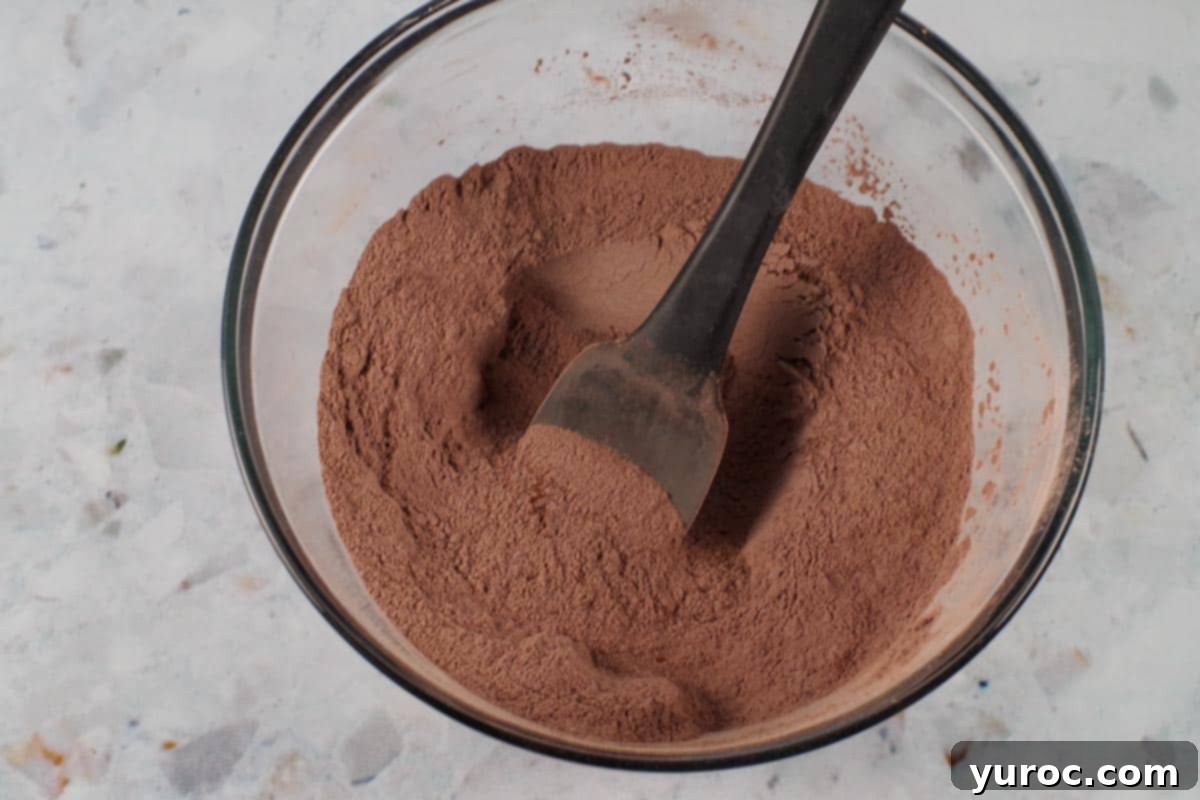 Blender Bliss Chocolate Zucchini Bread 7 Dry ingredients for the chocolate blender zucchini bread, including cocoa, flour, sugar, baking powder, baking soda, salt, and cinnamon, mixed together in a clear glass bowl with a black spatula.