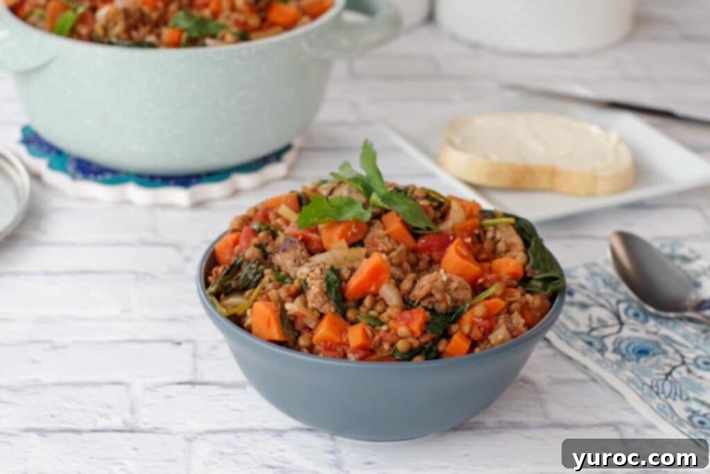 Lentil and sausage stew in blue bowl on white surface with light blue pot of stew in the background
