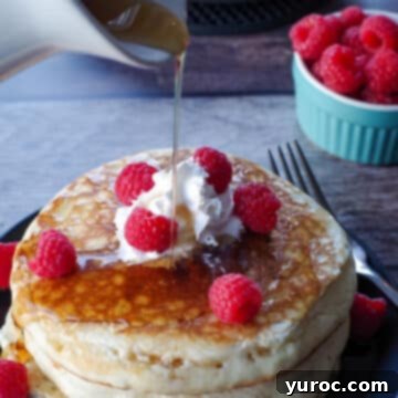 A stack of three fluffy pancakes, artfully garnished with fresh raspberries, a drizzle of syrup, and a dollop of whipped cream, presented on a black plate with more syrup being poured over them.