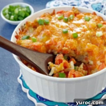 A comforting carrot casserole served in a white baking dish, with a wooden spoon scooping out a portion against a blue background, and dishes of green onions and oven mitts in the soft background.