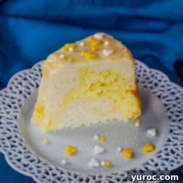 A delicate slice of lemon daffodil cake, with its distinct yellow and white layers, resting on a white plate with lattice trim, against a soft blue fabric background.