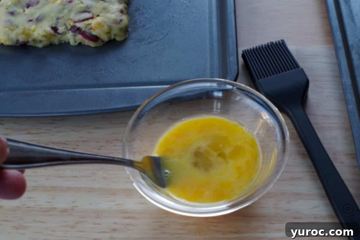 Egg mixed with water in a glass bowl, with biscotti loaves on a cookie sheet in the background, preparing for egg wash.