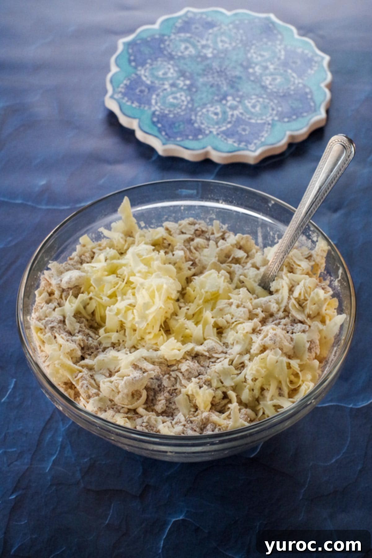 A bowl containing oats, flour, and brown sugar, with cubes of cold butter being cut into the dry ingredients using a pastry cutter, forming a crumbly mixture.