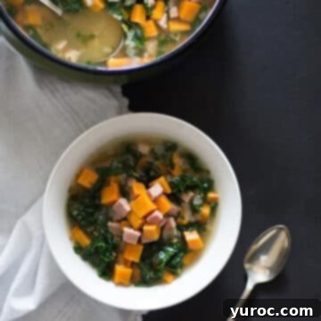 Sweet potato ham and kale soup in a white bowl with a spoon and a pot of soup behind