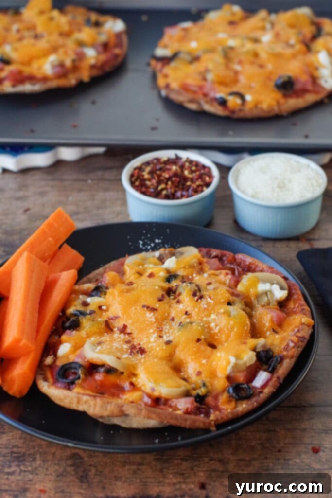 Two healthy pita pizzas on a black plate with carrot sticks, and more pizzas on a baking sheet in the background.