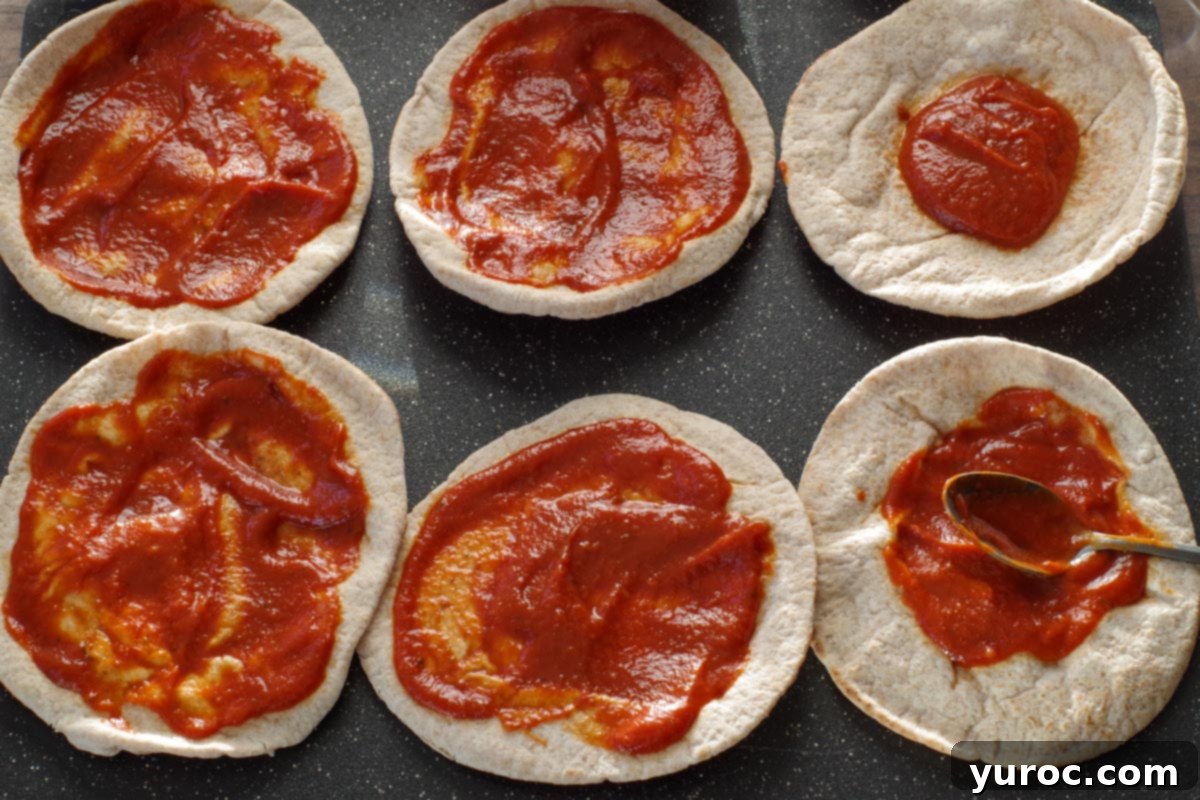 Six pitas on a cutting board with pizza sauce being spread on them, illustrating step 1.