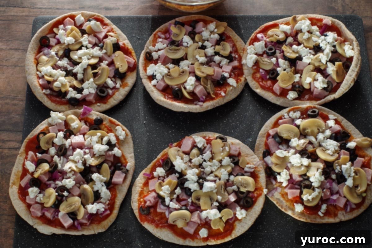 Mushrooms, black olives, feta cheese, and onion being sprinkled on pita pizzas, illustrating step 3.