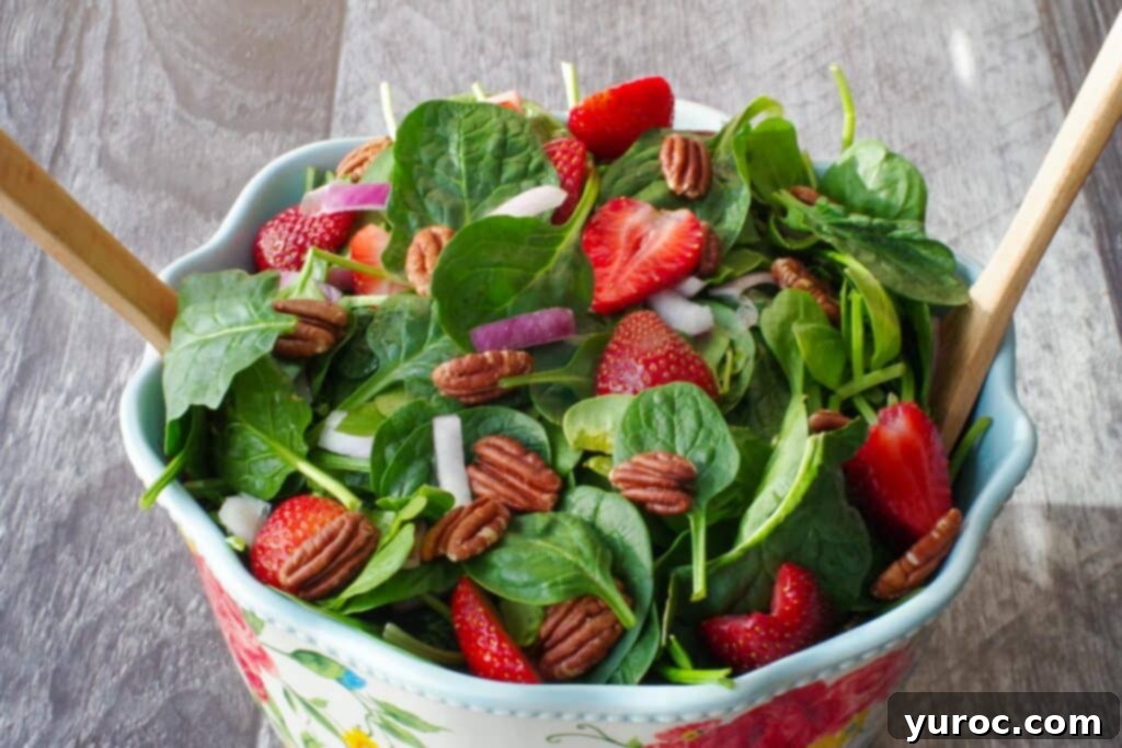 A vibrant strawberry spinach pecan salad, elegantly presented in a large floral-print bowl, accompanied by a pair of wooden serving utensils, showcasing a healthy and appealing side dish.
