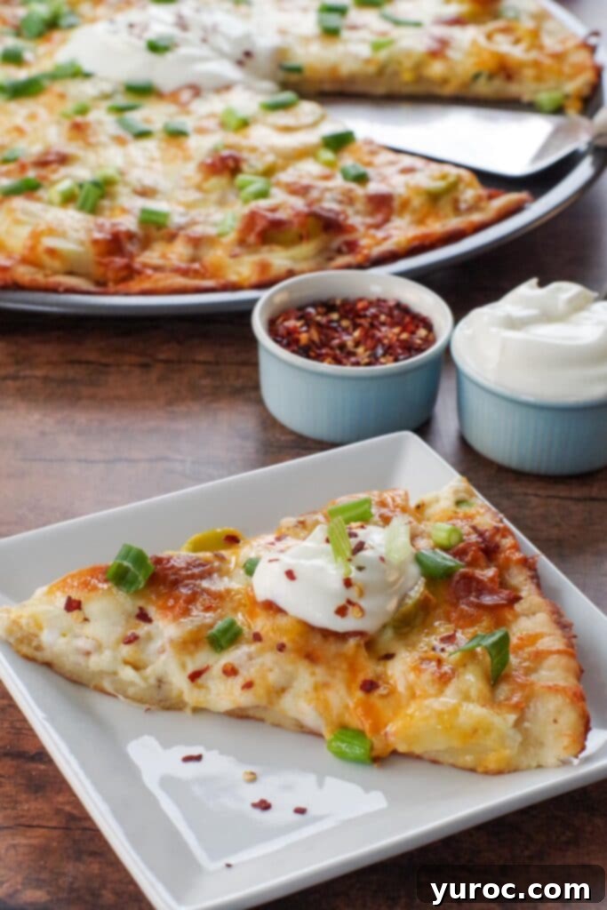 Another enticing view of a slice of Perogy Pizza on a white plate, with the whole pizza and small bowls of chili flakes and sour cream softly blurred in the background, inviting another taste.