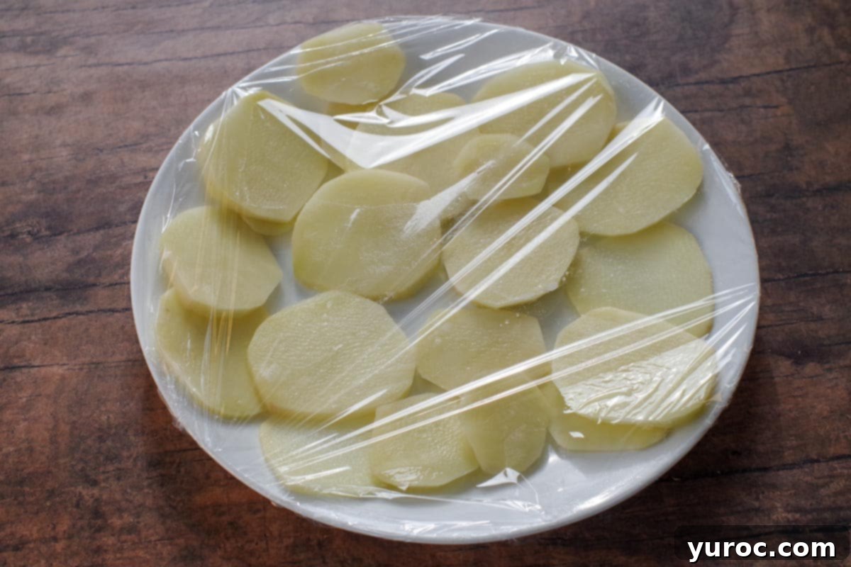 Delicate potato slices meticulously arranged on a pristine white plate, tightly sealed with clear plastic wrap, indicating preparation for microwave cooking.