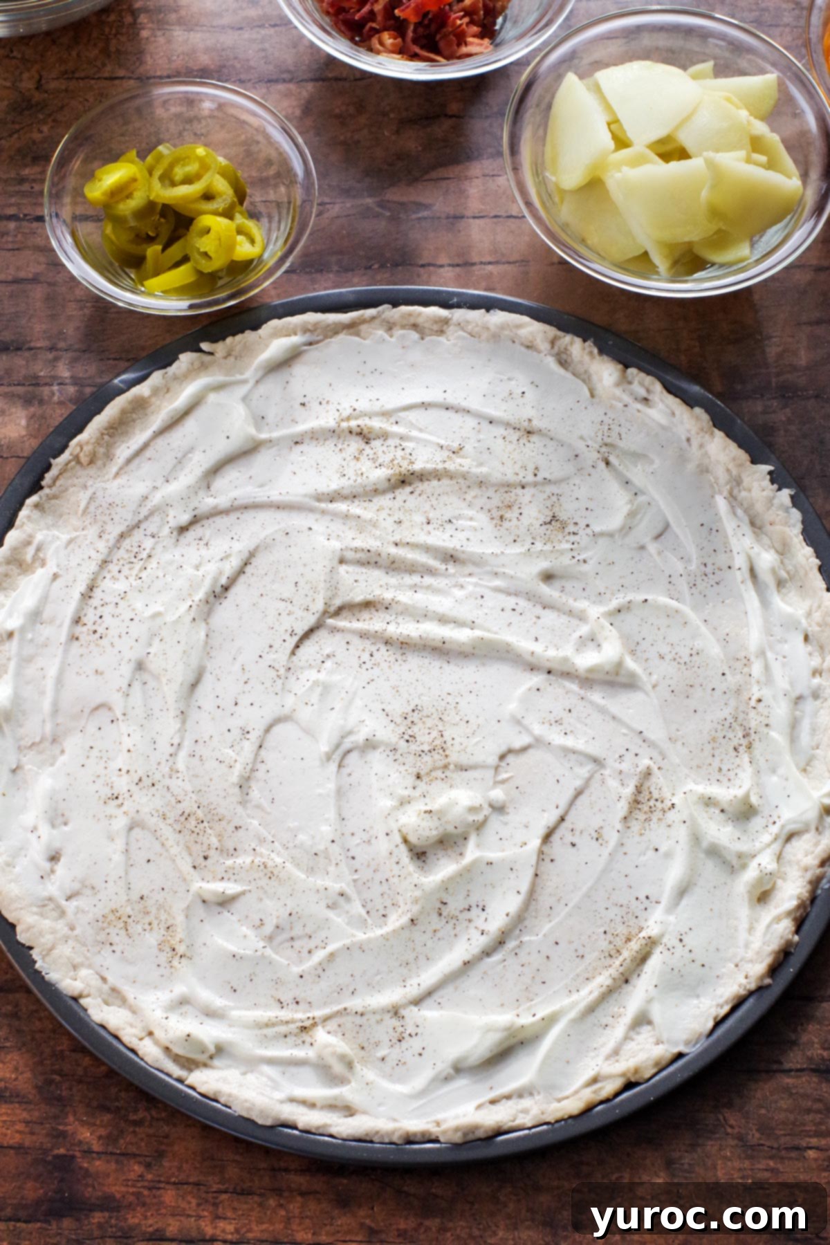 A pristine pizza dough base generously covered with a creamy layer of sour cream, seasoned with an even sprinkle of salt and black pepper, preparing for the next delicious layers.