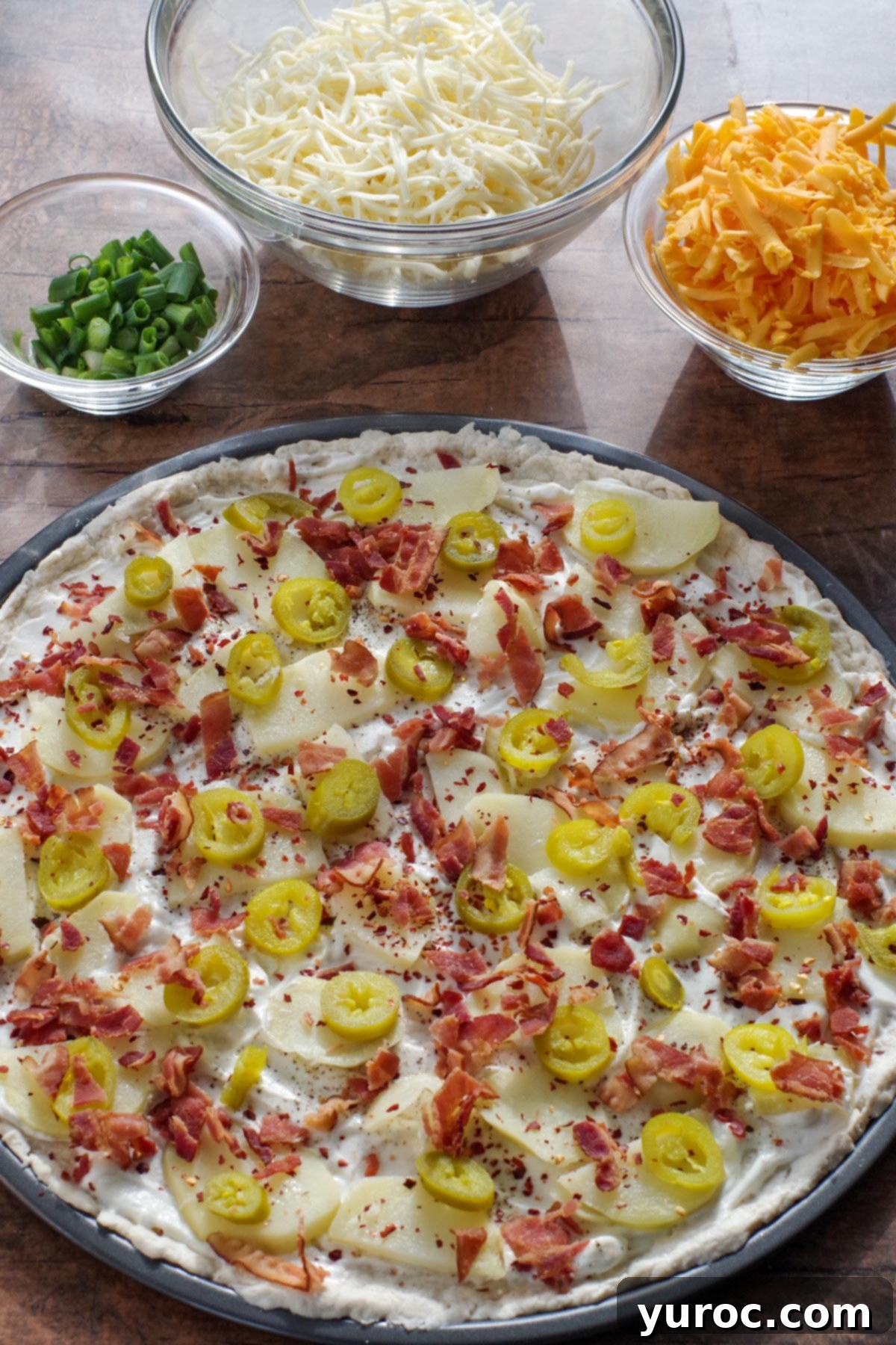 A pizza dough base layered with sour cream and chili flakes, now topped with perfectly arranged cactus-cut potatoes, vibrant jalapeños, and savory bacon pieces, awaiting the final cheese layer.