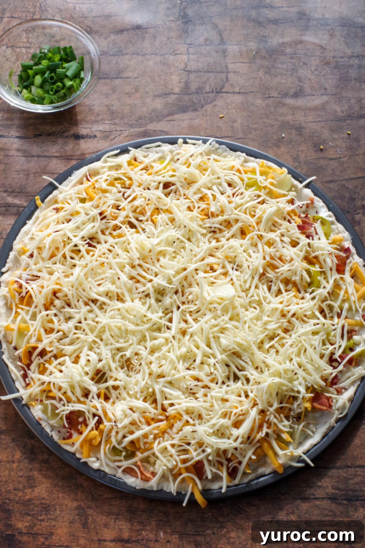 The pizza base, now fully loaded with all its savory toppings, is generously blanketed with a rich blend of shredded cheddar and mozzarella cheeses, ready for baking.