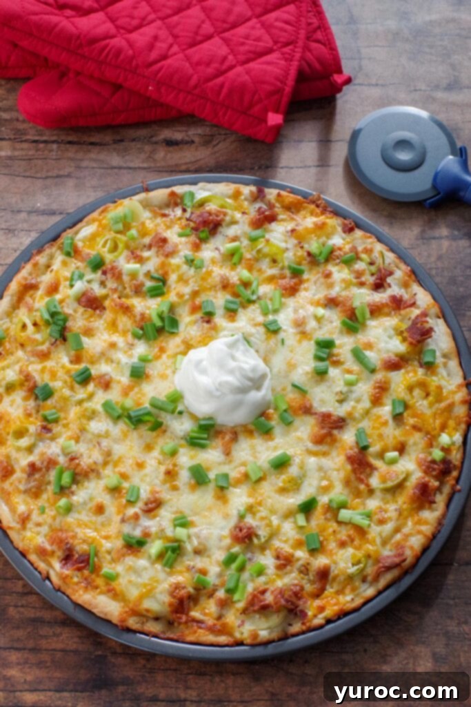 A perfectly baked, whole Perogy Pizza sits invitingly on a rustic brown surface, flanked by a pair of red oven mitts and a pizza cutter, signaling it's ready to be enjoyed.