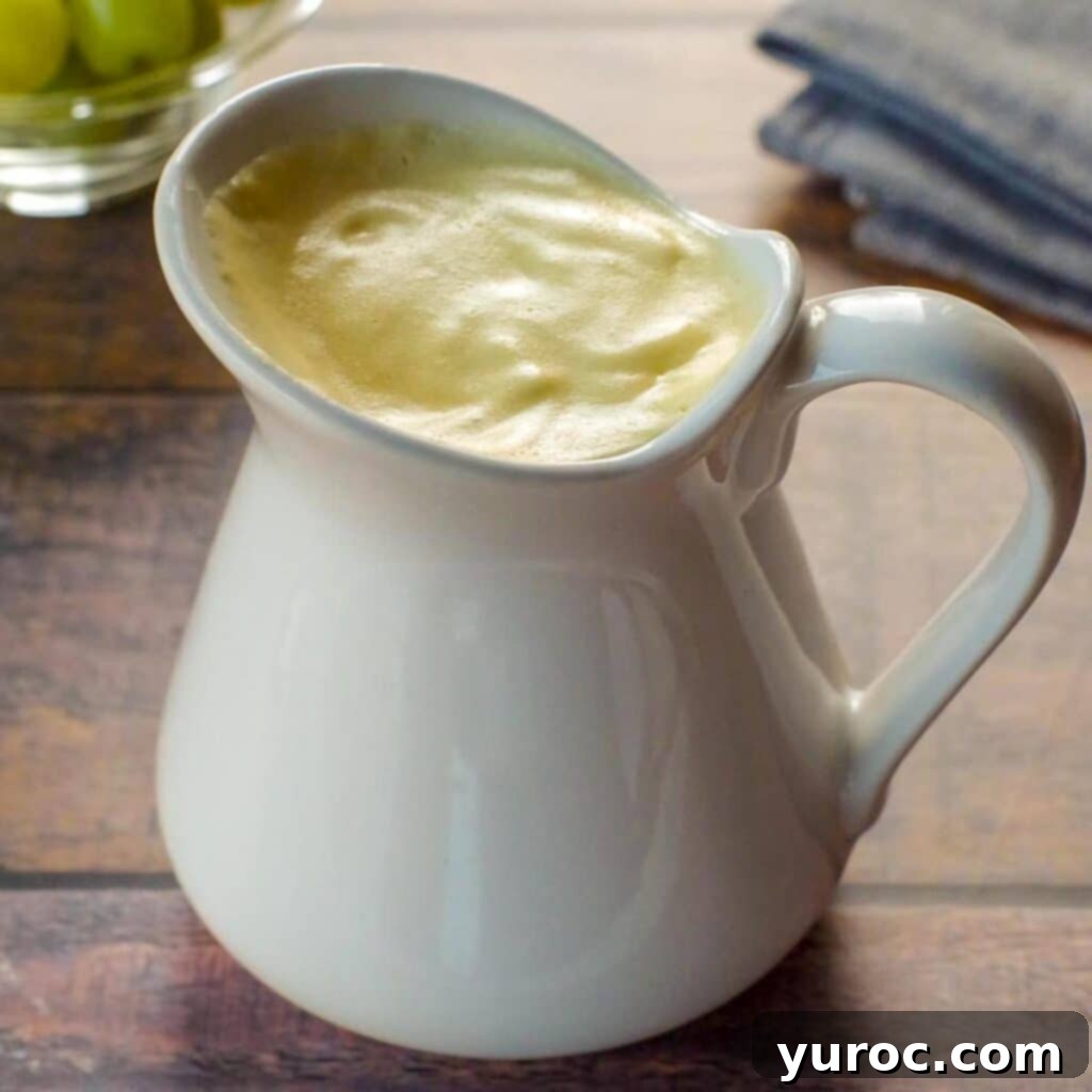 Healthy Hollandaise sauce in a large white creamer, with a bowl of green grapes in background and grey linen