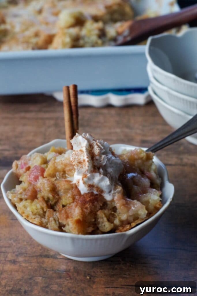 A close-up vertical shot of fresh rhubarb stalks in a charming white petal bowl, with a single cinnamon stick. The background is softly blurred, showing a full pan of baked sticky rhubarb pudding, highlighting the ingredients and the delicious final product.