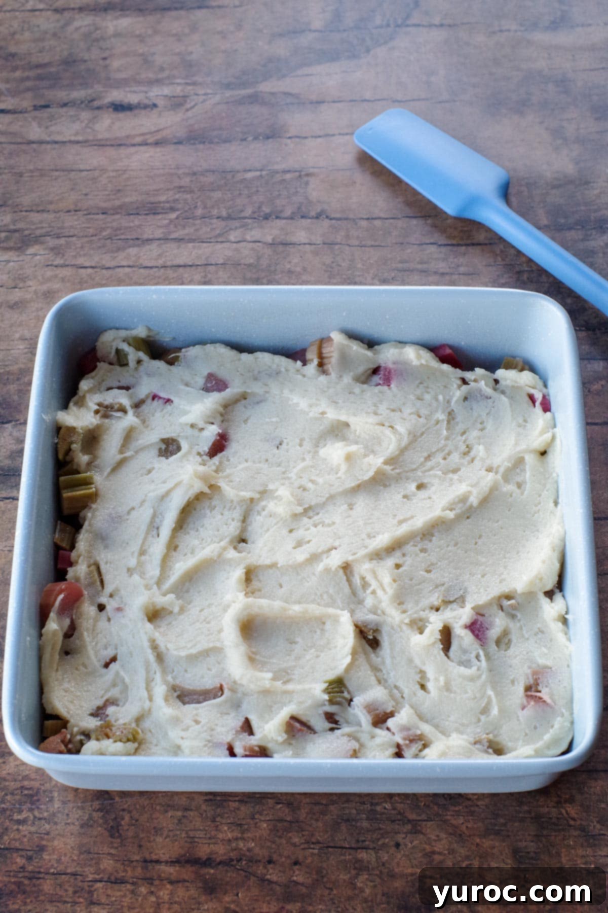 The smooth cake batter evenly spread over the layer of chopped rhubarb in the baking pan, forming the middle layer of the pudding.