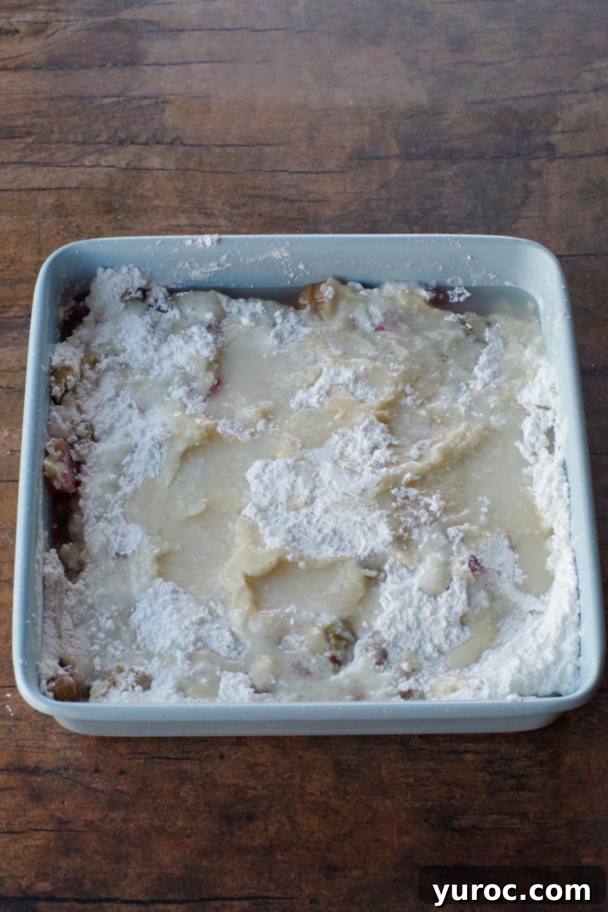 Water being carefully poured over the dry powdered sugar and cornstarch layer, which sits atop the cake batter and rhubarb, creating the final pre-bake layer.