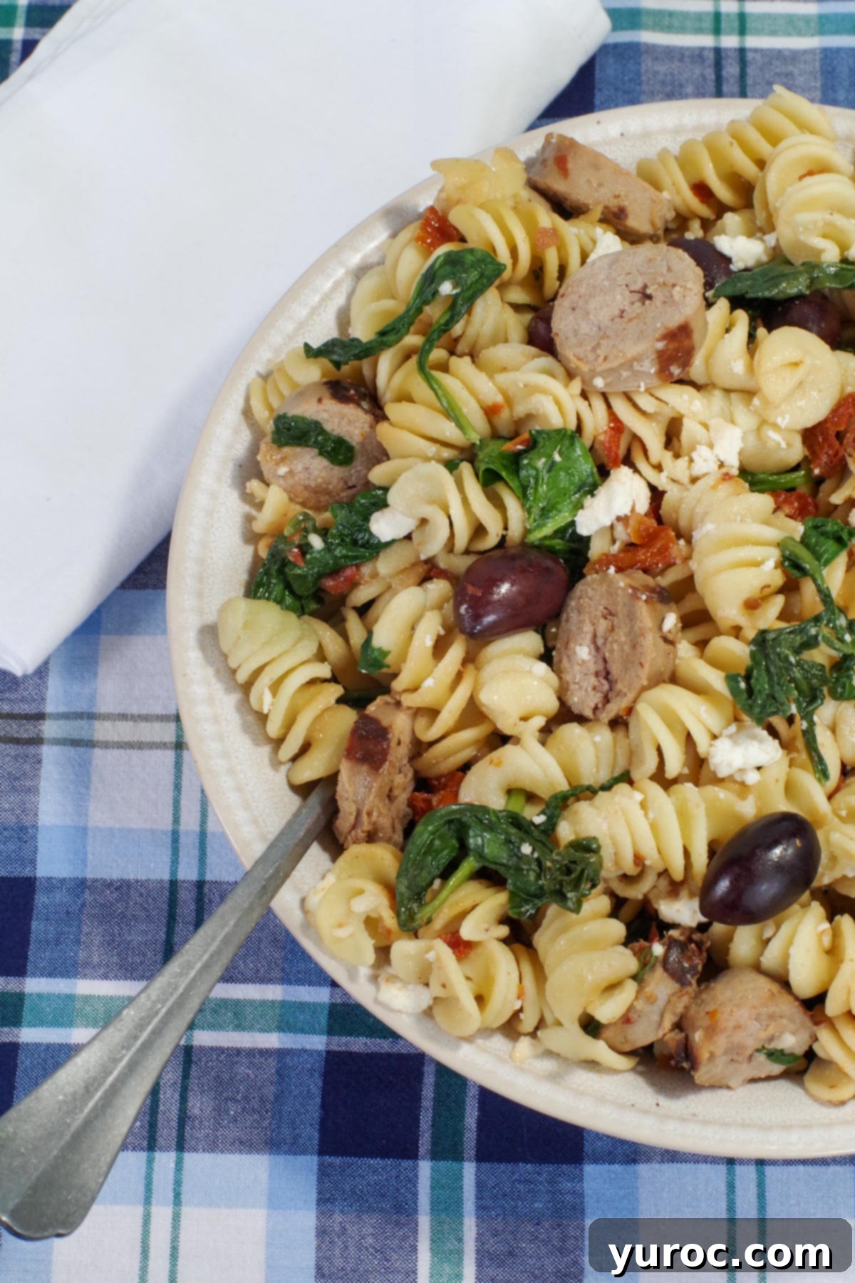 Sun-Kissed Aegean Pasta 10 Close-up of Mediterranean pasta in a blue bowl with a wooden spoon, garnished with fresh herbs, against a white napkin.