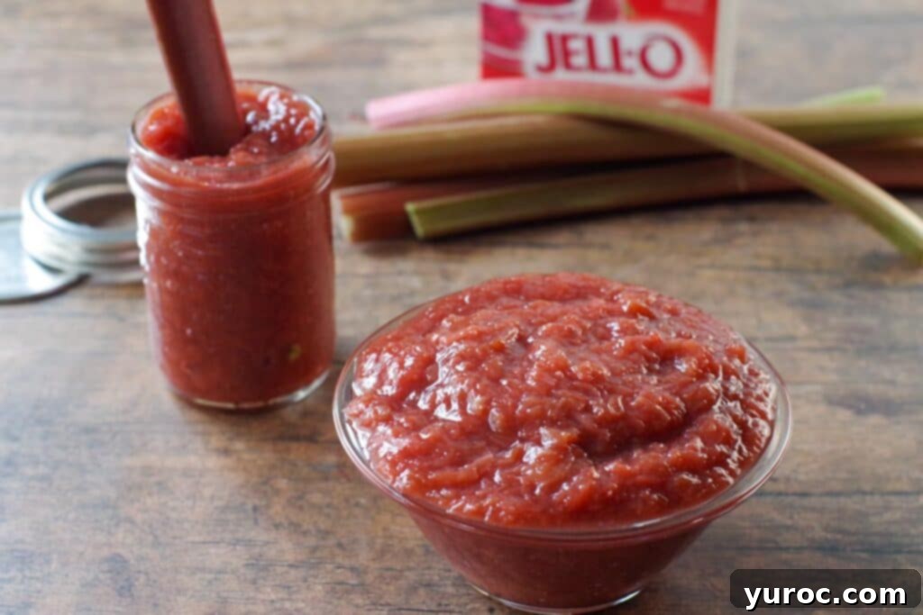 Freshly made stewed rhubarb in a glass bowl, with a jar of prepared rhubarb, fresh rhubarb stalks, and a box of strawberry gelatin in the background, showcasing the key ingredients and delicious result.