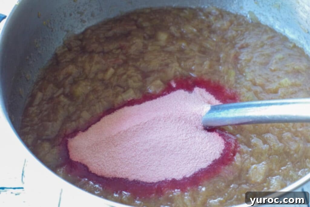 Dry strawberry gelatin powder being added to the hot stewed rhubarb mixture in a saucepan, ready to be stirred in.