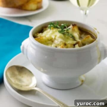 Classic French Onion Soup in a white handled bowl, served on a white plate with a small spoon, showcasing its melted Gruyere topping.