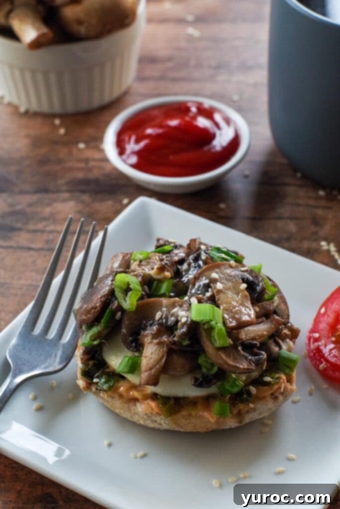A stack of two open-faced Breakfast Mushroom Melts on English muffins, with melted mozzarella, egg, and mushrooms, garnished with fresh green onion and sesame seeds. Sliced tomatoes and a small dish of ketchup are on the side.