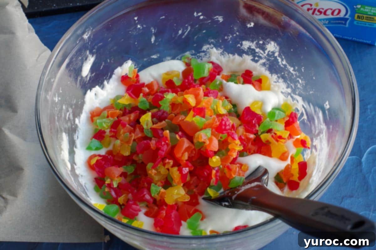 chopped jujubes added to large glass bowl with melted marshmallows, white chocolate chips and butter, with a black spatula in it