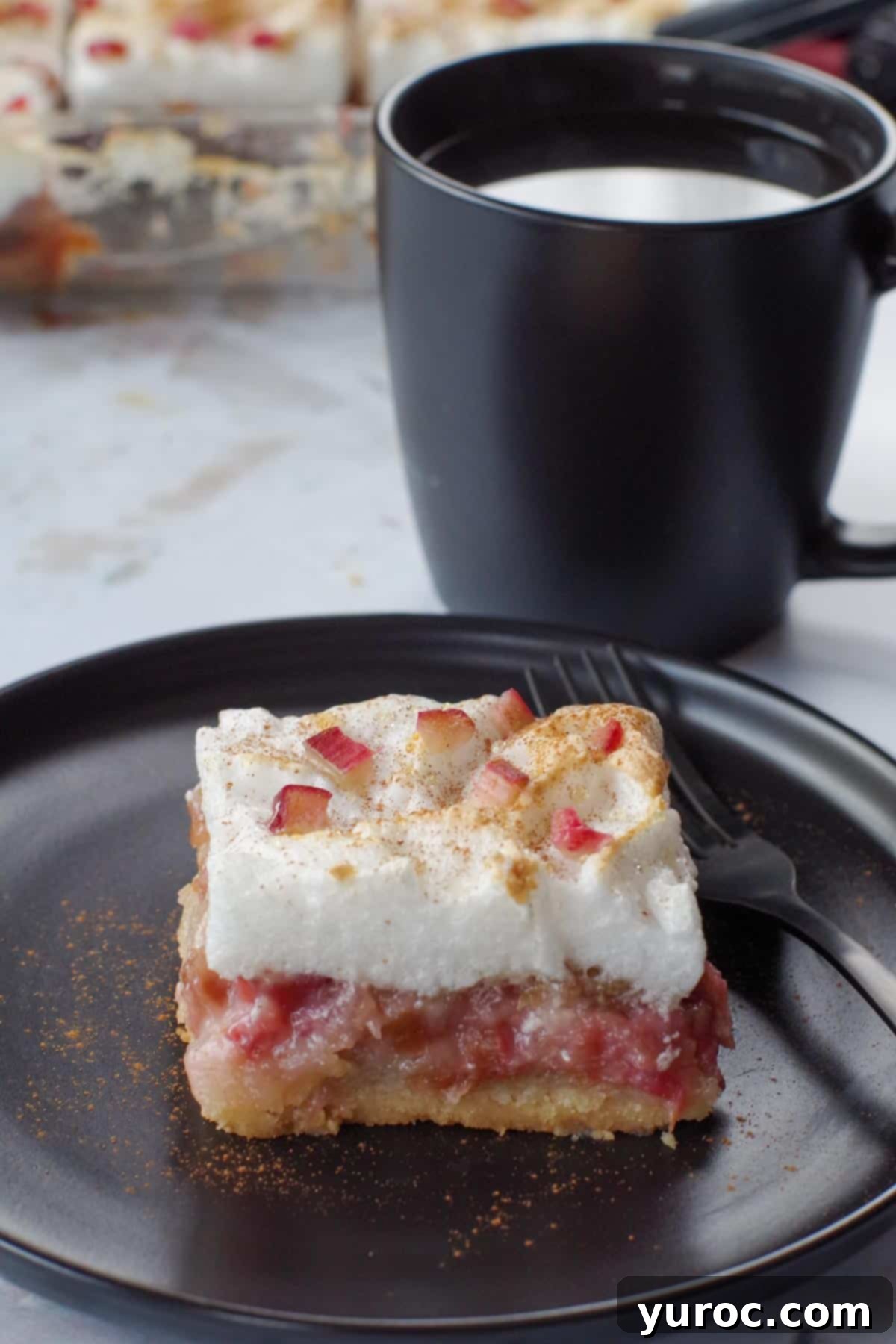 a piece of rhubarb meringue torte on a black plate with a fork and a black cup of coffee in background