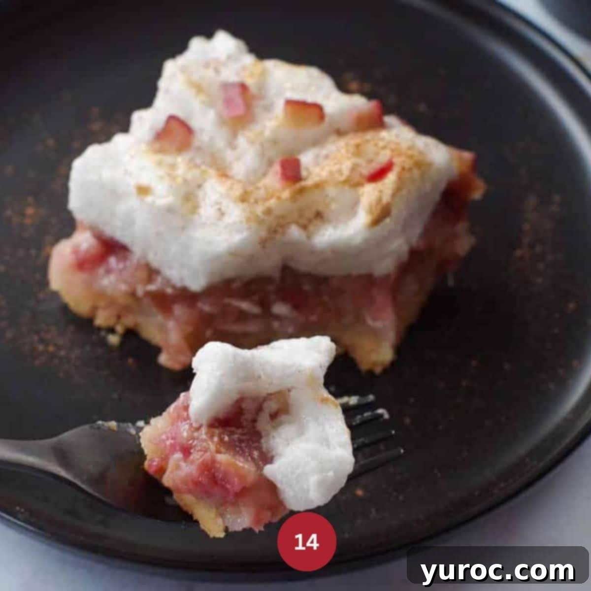 A piece of Rhubarb Meringue Torte garnished with rhubarb and cinnamon, on a black plate,  with a fork cutting into it.
