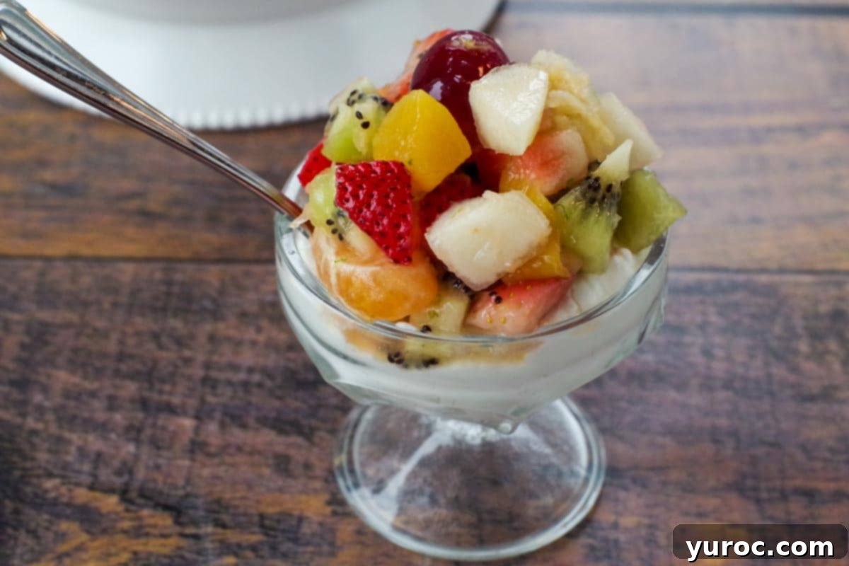 fruit salad over Greek yogurt in a glass dessert dish, with a spoon (on faux wooden surface). A light and healthy fruit salad, perfect alongside a hearty breakfast.