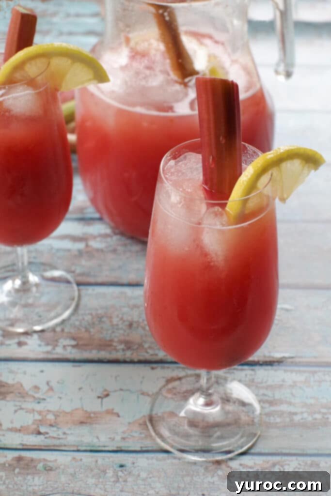 Effortless Rhubarb Lemonade 1 A glass of vibrant pink rhubarb lemonade, with a matching pitcher and another glass in the soft-focus background, suggesting a delightful summer refreshment.