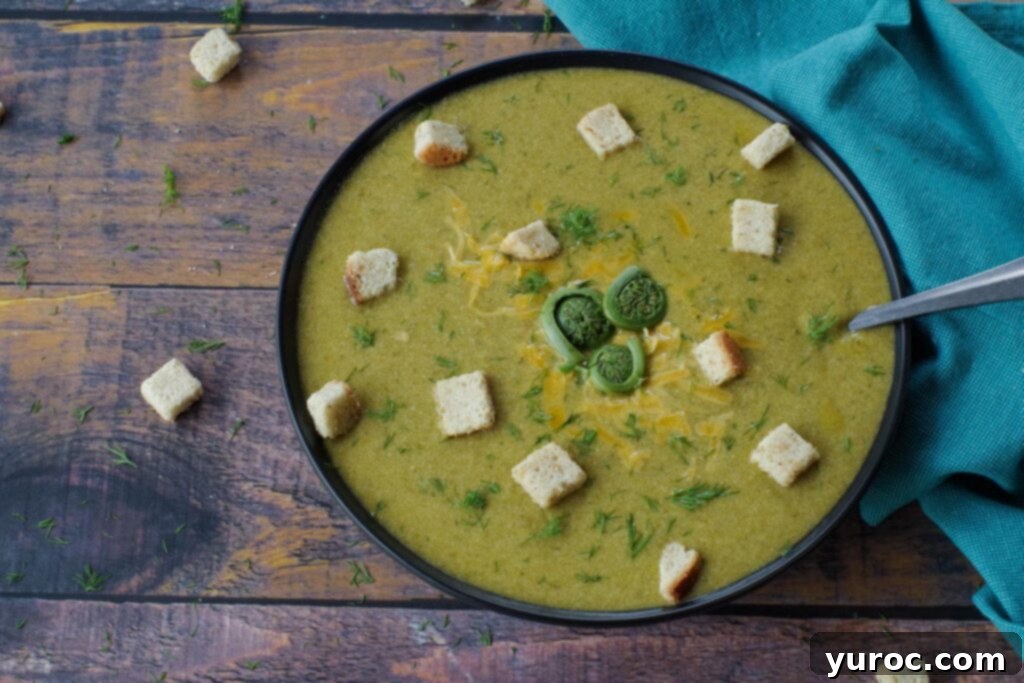 A bowl of creamy fiddlehead soup garnished with croutons, fresh dill, and whole fiddleheads, with a spoon beside it.