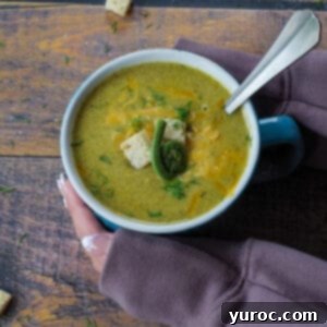 A close-up of a mug filled with creamy fiddlehead soup, adorned with fresh dill and a spoon, held by hands for warmth.