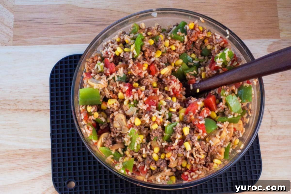 Green peppers, onions, rice, ground beef, ketchup, garlic and canned corn in a large glass bowl with wooden spoon.
