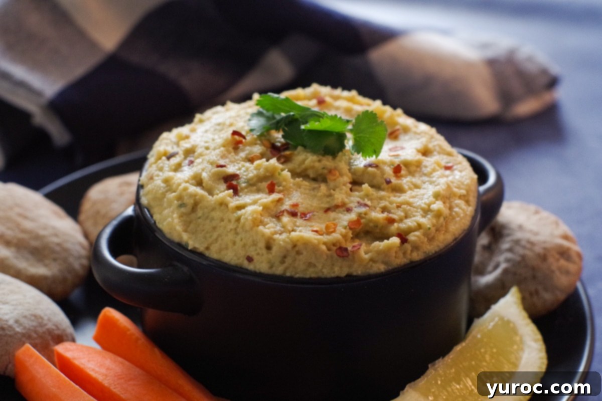 Light Yogurt Hummus 2 Creamy yogurt hummus in a black bowl, garnished with fresh cilantro and a sprinkle of red pepper flakes, surrounded by an assortment of colorful fresh vegetables and mini pita breads on a black plate.