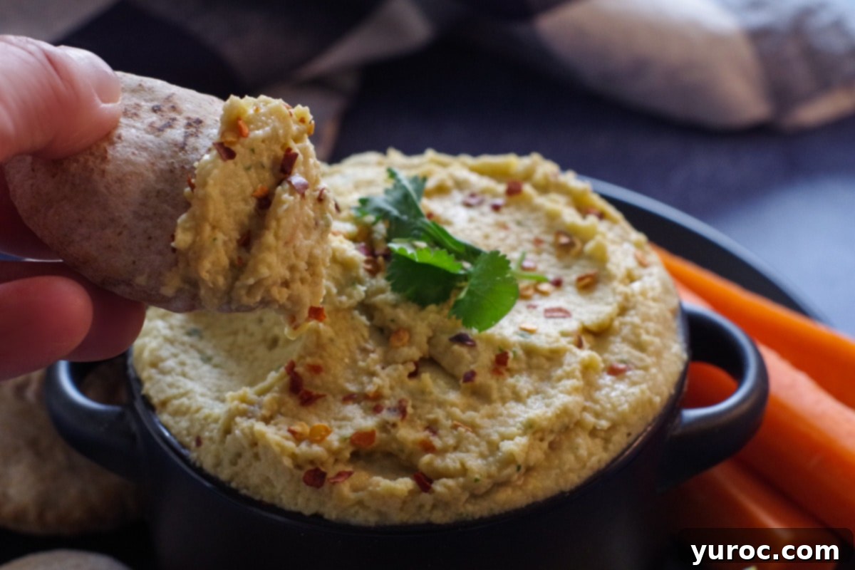 A mini pita bread is gently dipped into the creamy yogurt hummus, showcasing its delightful texture, with more fresh vegetables and mini pitas artfully arranged on a black plate in the background.
