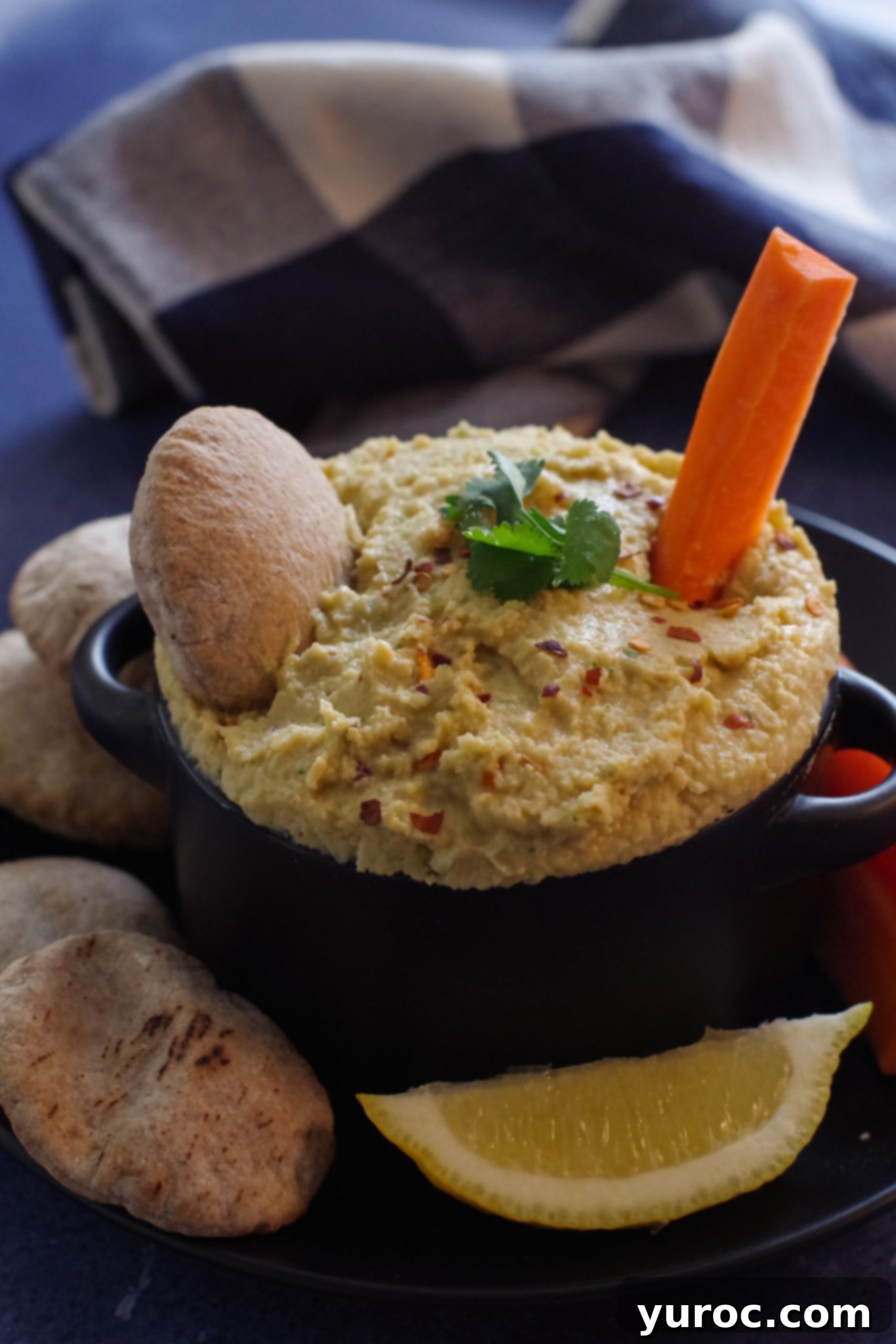 A black bowl filled with creamy yogurt hummus, garnished with cilantro and red pepper flakes, holds a mini pita and a carrot stick. The bowl rests on a black plate, surrounded by an array of fresh vegetables and more mini pita breads.