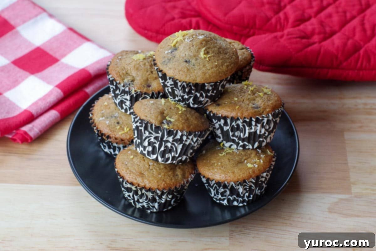 Heavenly Saskatoon Berry Muffins 12 Saskatoon berry muffins piled up on a black plate with red oven mitts and a red and white checkered tablecloth in the background