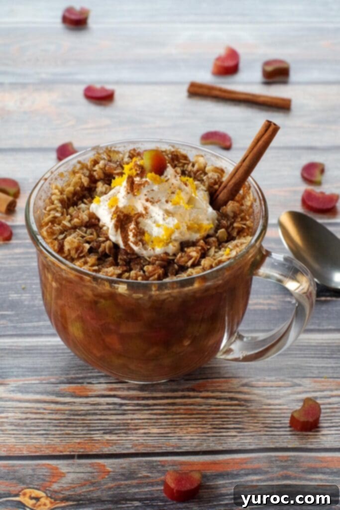 Close-up of a microwave rhubarb crisp in a mug, generously topped with fluffy whipped cream, adorned with a cinnamon stick, and accompanied by a spoon, with scattered fresh rhubarb pieces and cinnamon sticks on the side for garnish.