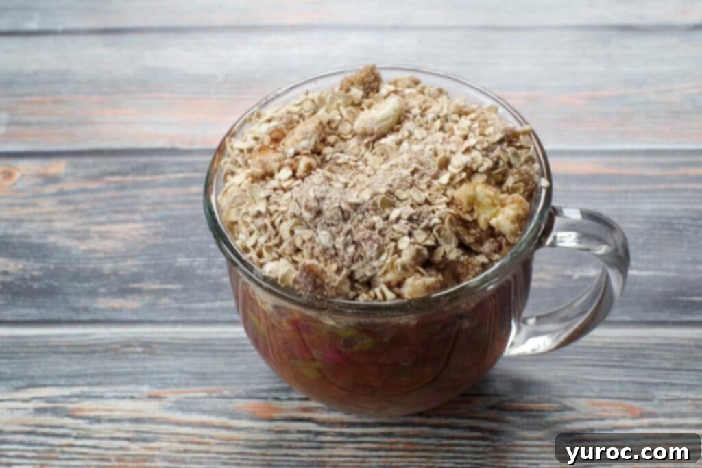 The prepared crumbly crisp topping generously spooned over the vibrant rhubarb filling in a glass mug, perfectly set up for microwaving.