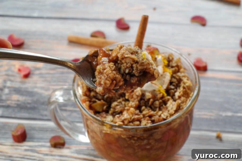 A delectable microwave rhubarb crisp served in a mug, garnished with a swirl of whipped cream and a cinnamon stick. A spoon holds a perfect bite of the warm crisp, with fresh rhubarb and cinnamon sticks artfully scattered on a rustic wooden surface in the background.