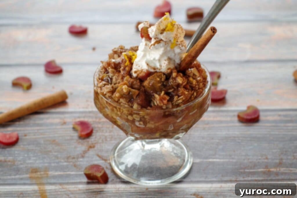 A beautifully presented rhubarb crisp in a clear glass dessert dish, artfully topped with fluffy whipped cream and a cinnamon stick. Scattered chopped rhubarb and cinnamon sticks in the background add to the visual appeal.