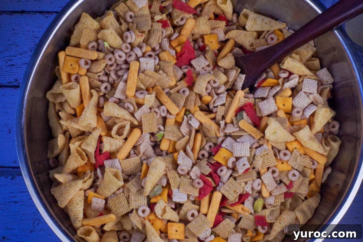 A large stainless steel bowl filled with a mix of Cheerios, Life cereal, Chex cereal, Bugles, pistachios, cheese nips, cheese bits, and red tortilla chips, before seasoning.