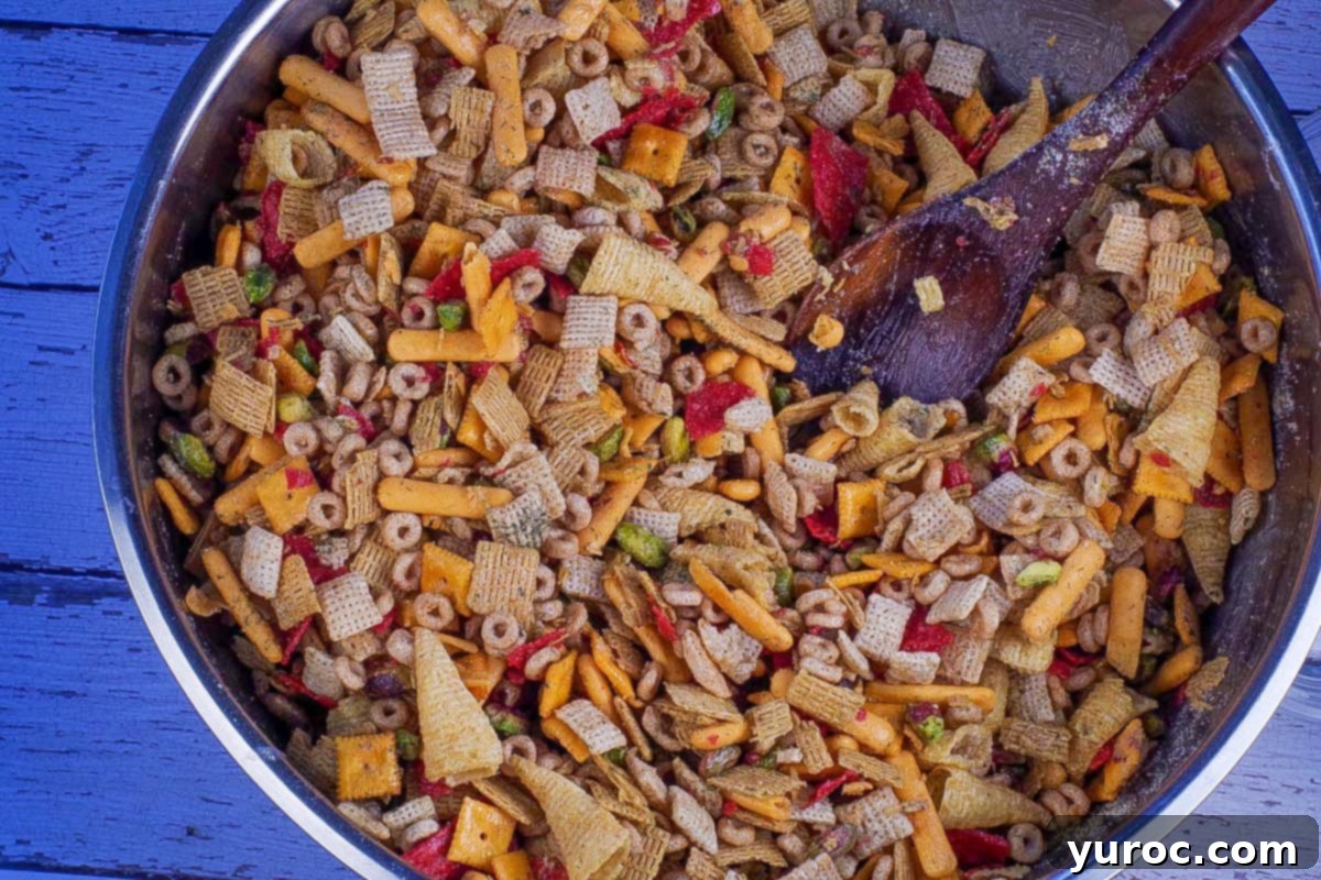 The butter and seasoning mixture being poured over the dry cereal and cracker blend in a large stainless steel bowl, with a wooden spoon for stirring.