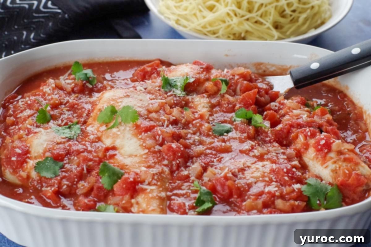 A dish of baked chicken cacciatore in a white casserole dish, with a black serving utensil and a bowl of noodles in the blurred background, suggesting a complete meal.