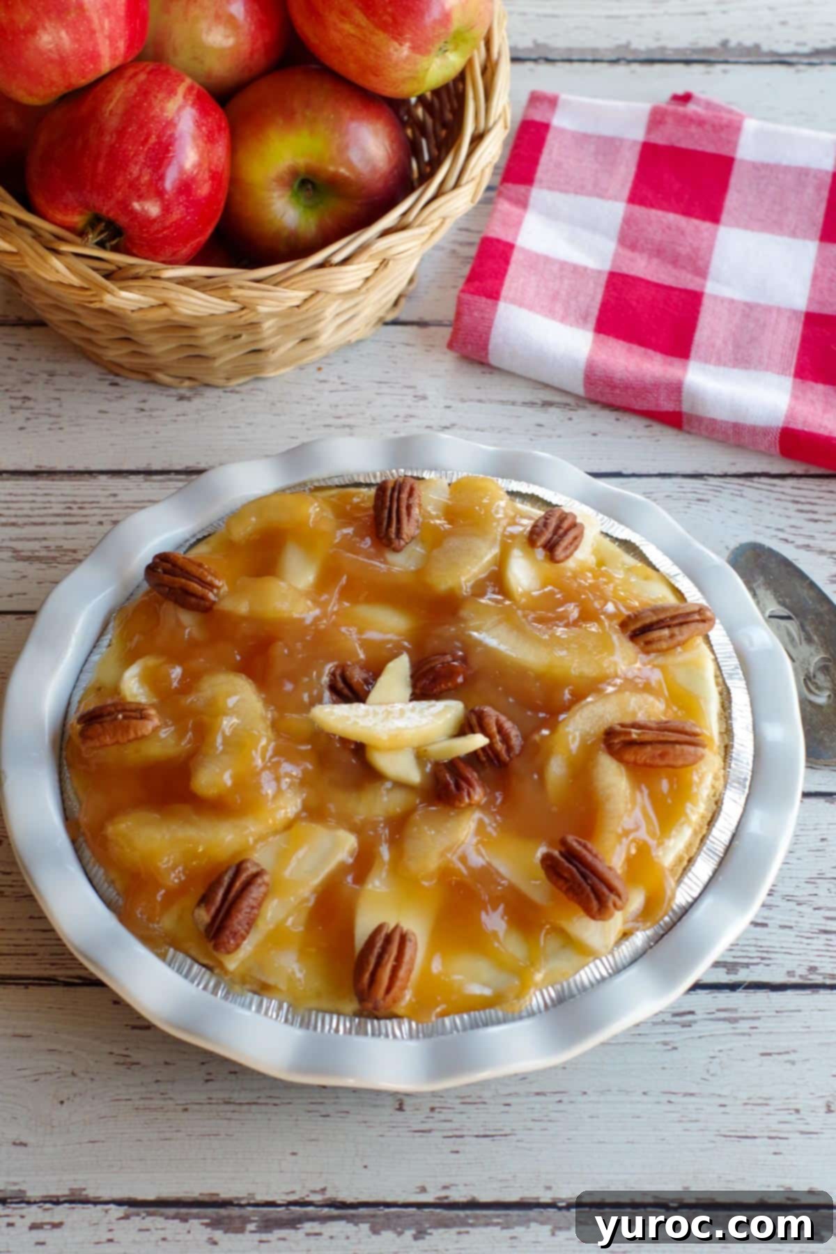 A whole, beautifully presented caramel apple cheesecake pie on a white wooden background, with a red checkered cloth behind it, ready to be served or stored.