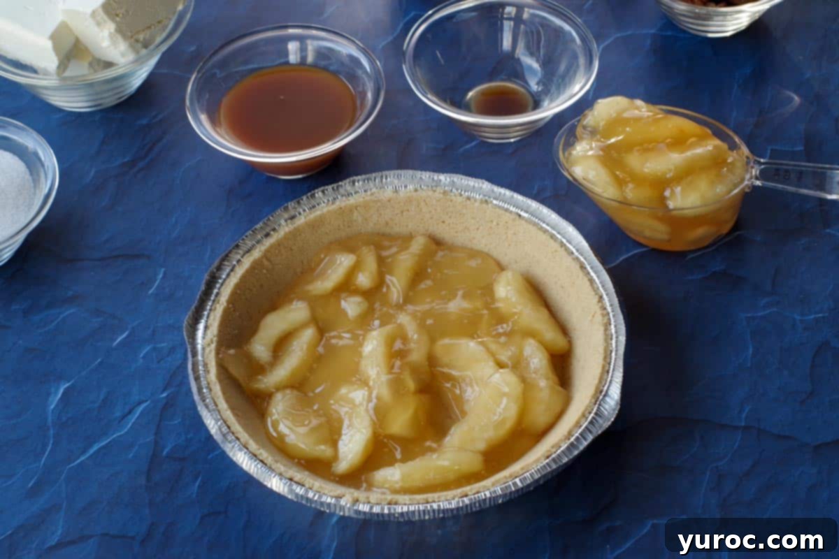 Apple pie filling being spooned evenly onto a graham cracker pie crust, creating the first layer of the cheesecake pie.