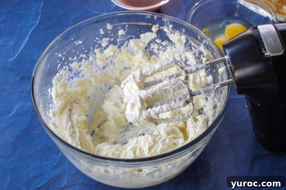 Cream cheese, sugar, and vanilla extract being beaten together in a large glass bowl with an electric hand mixer, creating a smooth cheesecake batter.
