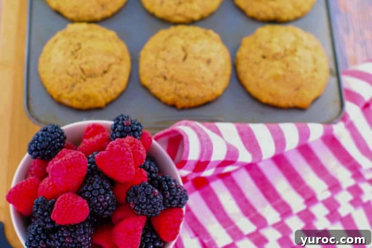 6 baked Old Fashioned Wheat bran muffins in a muffin tin, with a bowl of berries in front of it, as well as a red striped tea towel.